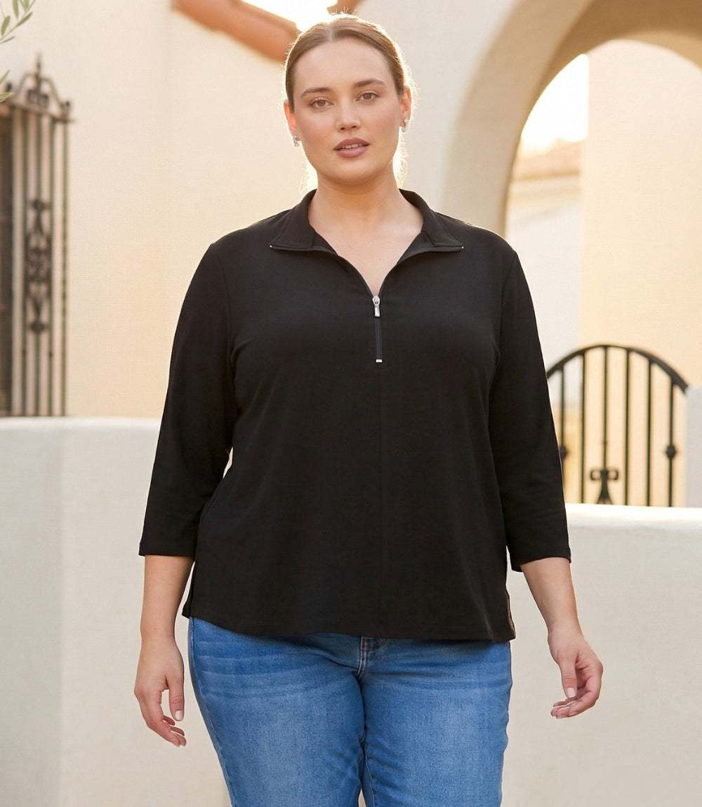 A woman wearing a black three-quarter sleeve mock-neck top with a short silver zipper at the collar, paired with blue jeans, standing in front of a beige stucco wall with an arched doorway.