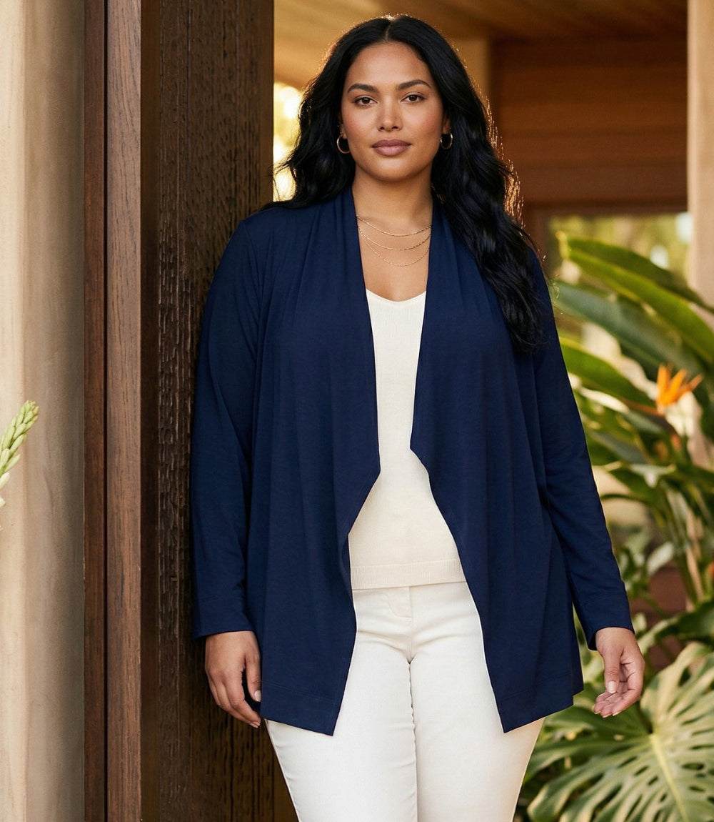 A woman modeling a navy blue open-front cardigan over a white top and white pants, standing in a warm-toned indoor setting with plants visible in the background.