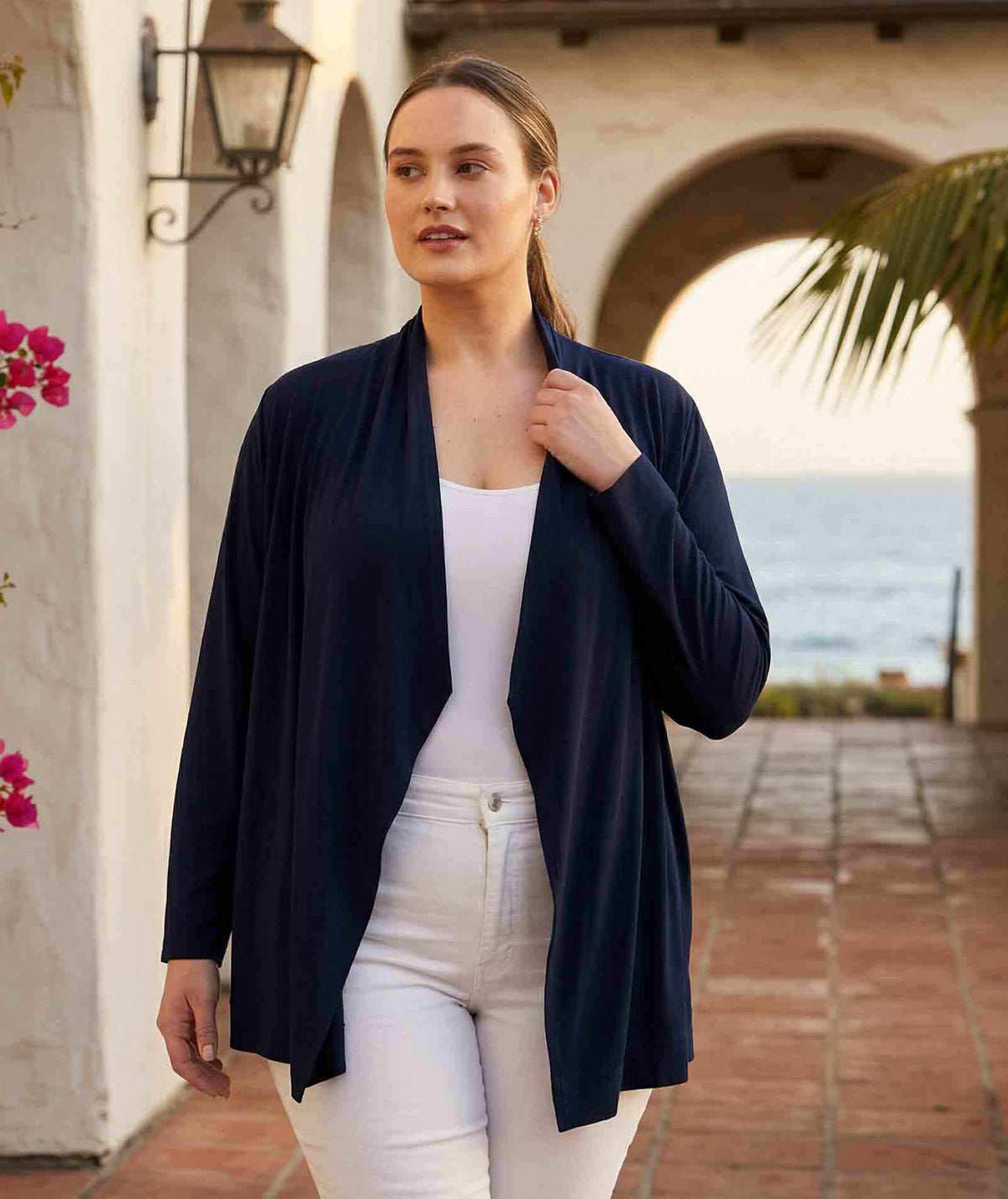Woman wearing a navy cardigan over a white top and pants, standing in a scenic location with ocean view.
