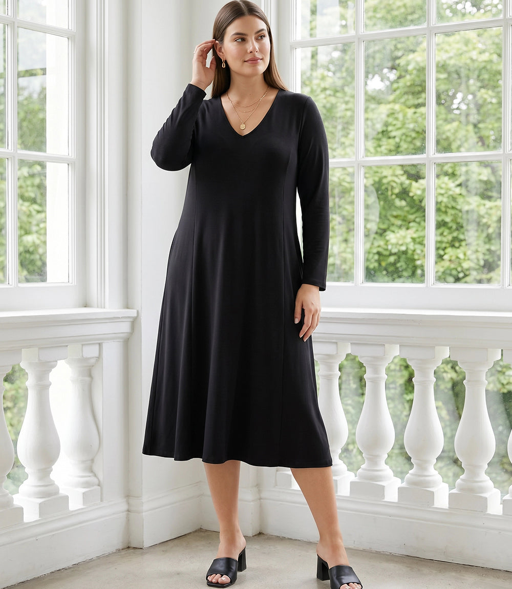 A woman modeling a long-sleeve black midi dress with a V-neckline, standing on a white balcony with large windows and green foliage visible in the background.