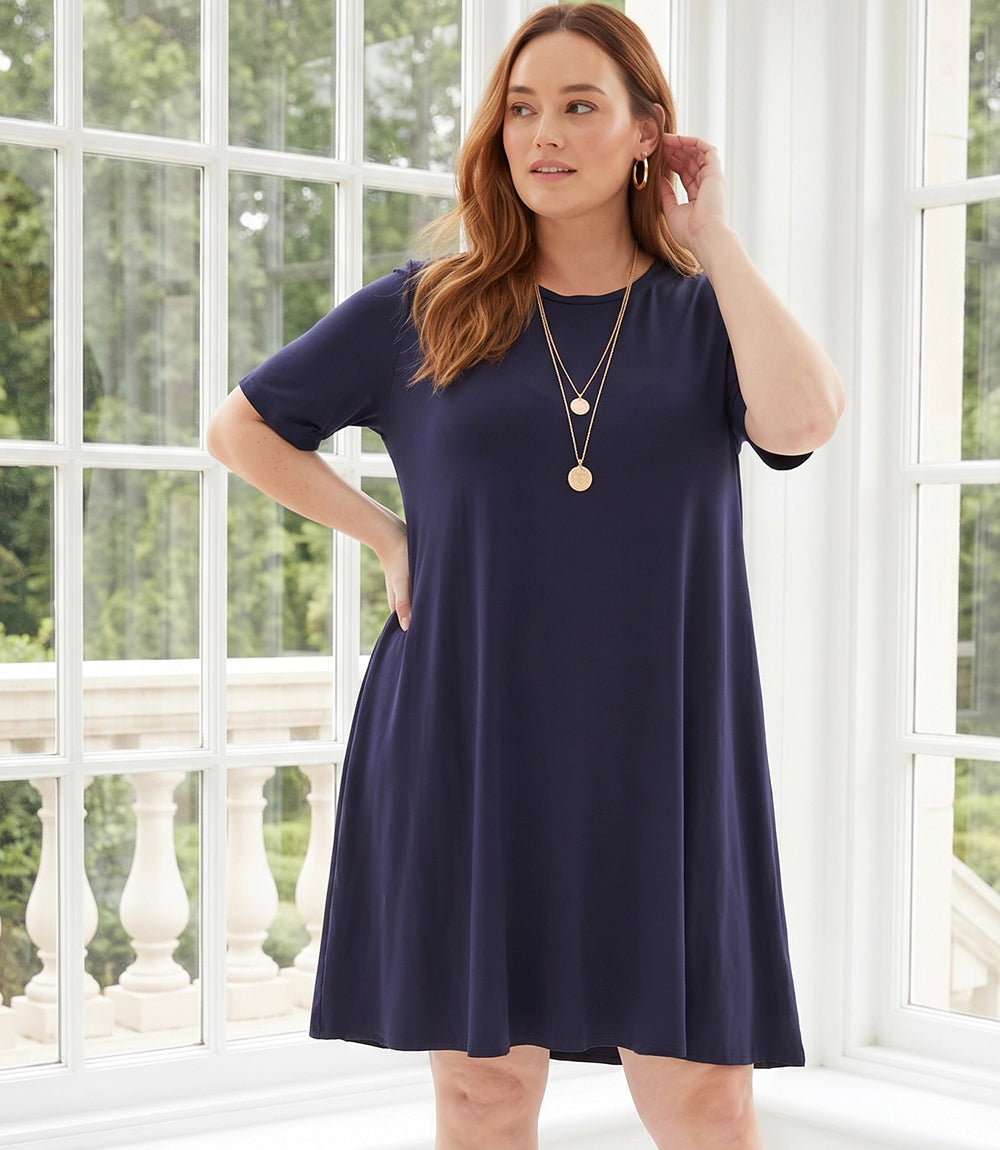 A woman modeling a short-sleeve navy blue shift dress with a round neckline, standing in front of a large white-framed window with outdoor greenery visible.