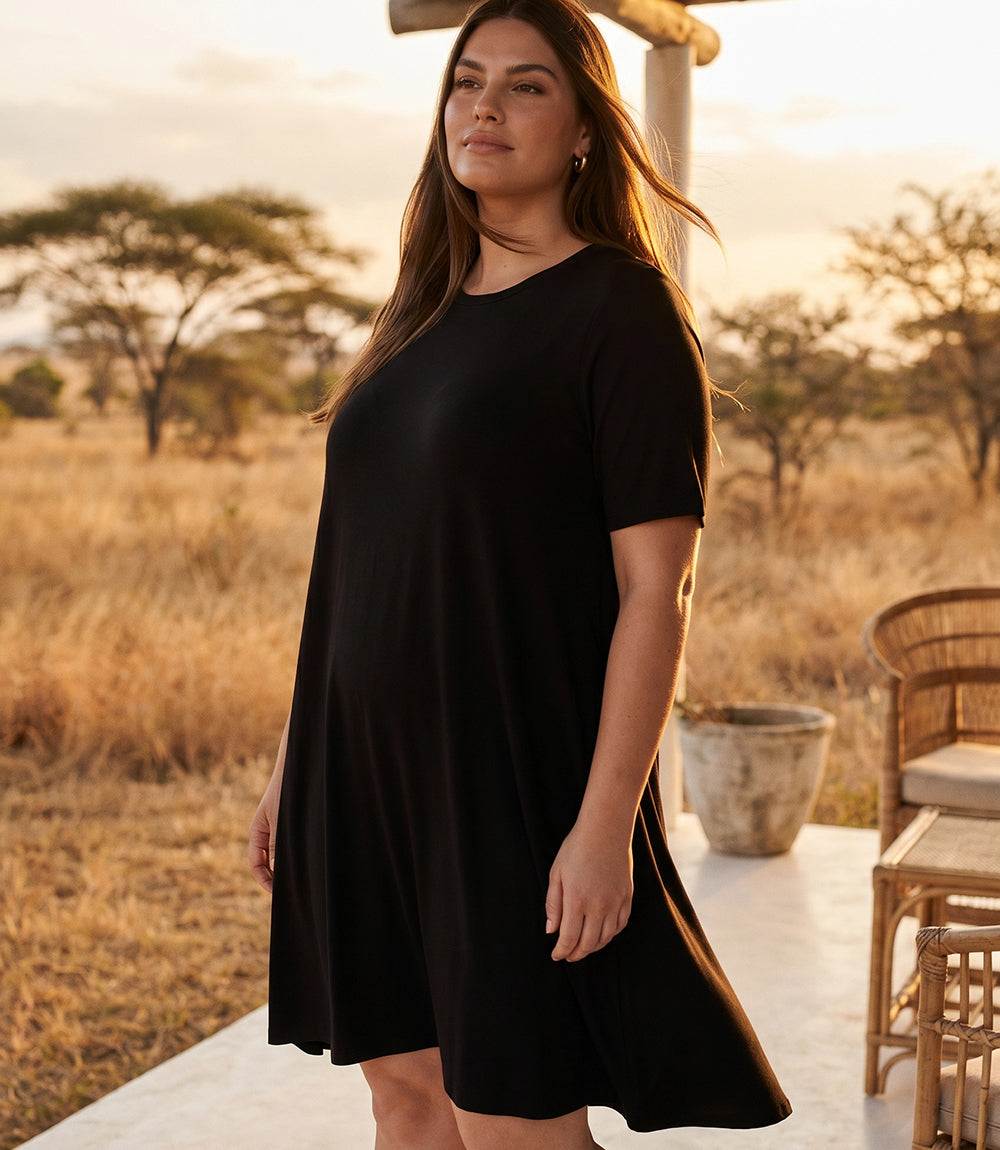 A woman wearing a black short-sleeve tunic dress stands on an outdoor patio at sunset with dry grass and trees in the background.