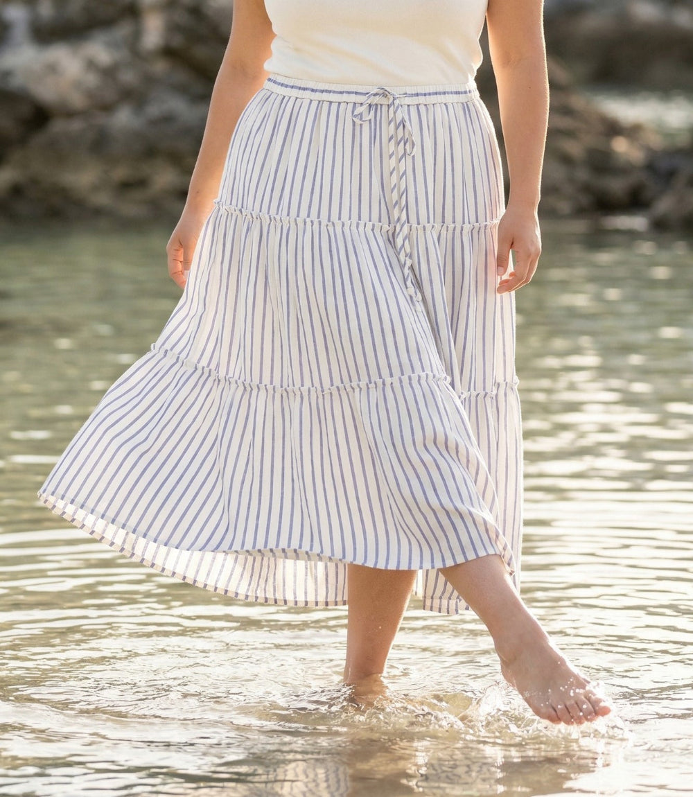 A woman walking barefoot in shallow water wearing a white tiered midi skirt with thin blue vertical stripes and a matching drawstring waist.