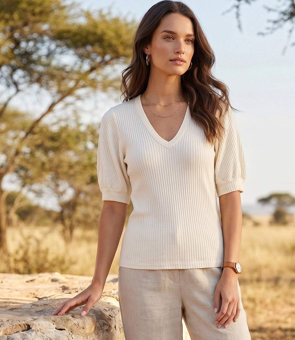 A woman stands outdoors in a sunlit savanna-like setting wearing a ribbed short-sleeve V-neck top in warm off-white and light beige trousers.
