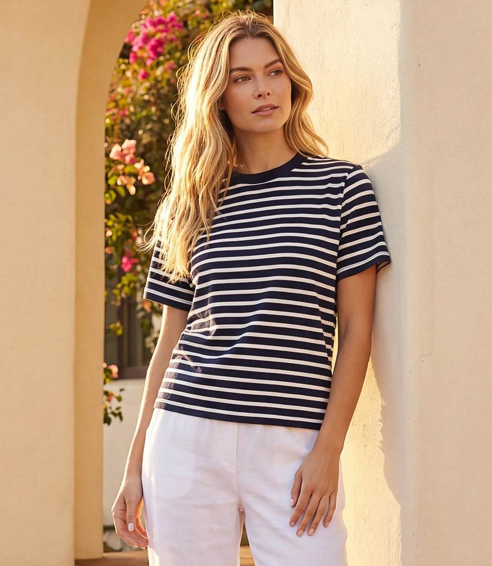 A woman wearing a navy and white horizontally striped short-sleeve t-shirt and white pants, leaning against a beige stucco wall in a sunlit outdoor setting.