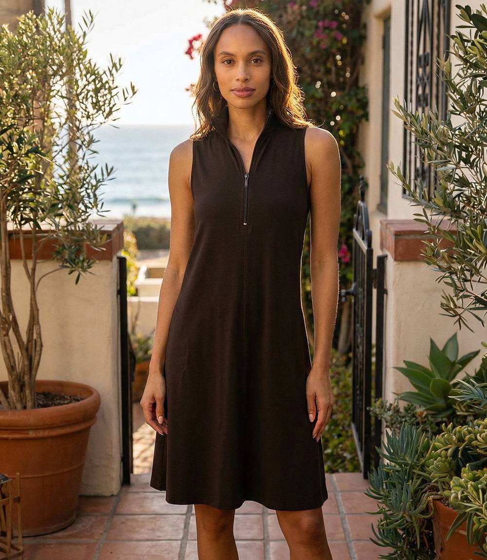 Woman wearing a black sleeveless dress standing outdoors with plants and ocean view.