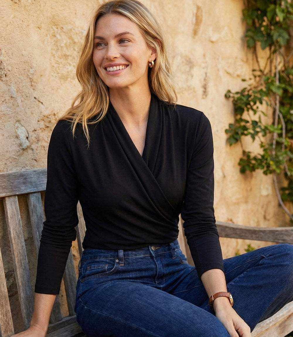 A woman sitting on a wooden bench wearing a black long-sleeve wrap-style top and blue jeans, with a warm stone wall and greenery in the background.