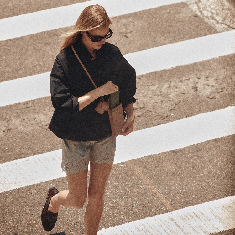 A woman walking across a crosswalk wearing a black button-up shirt, light gray shorts, black loafers, and carrying a brown shoulder bag.