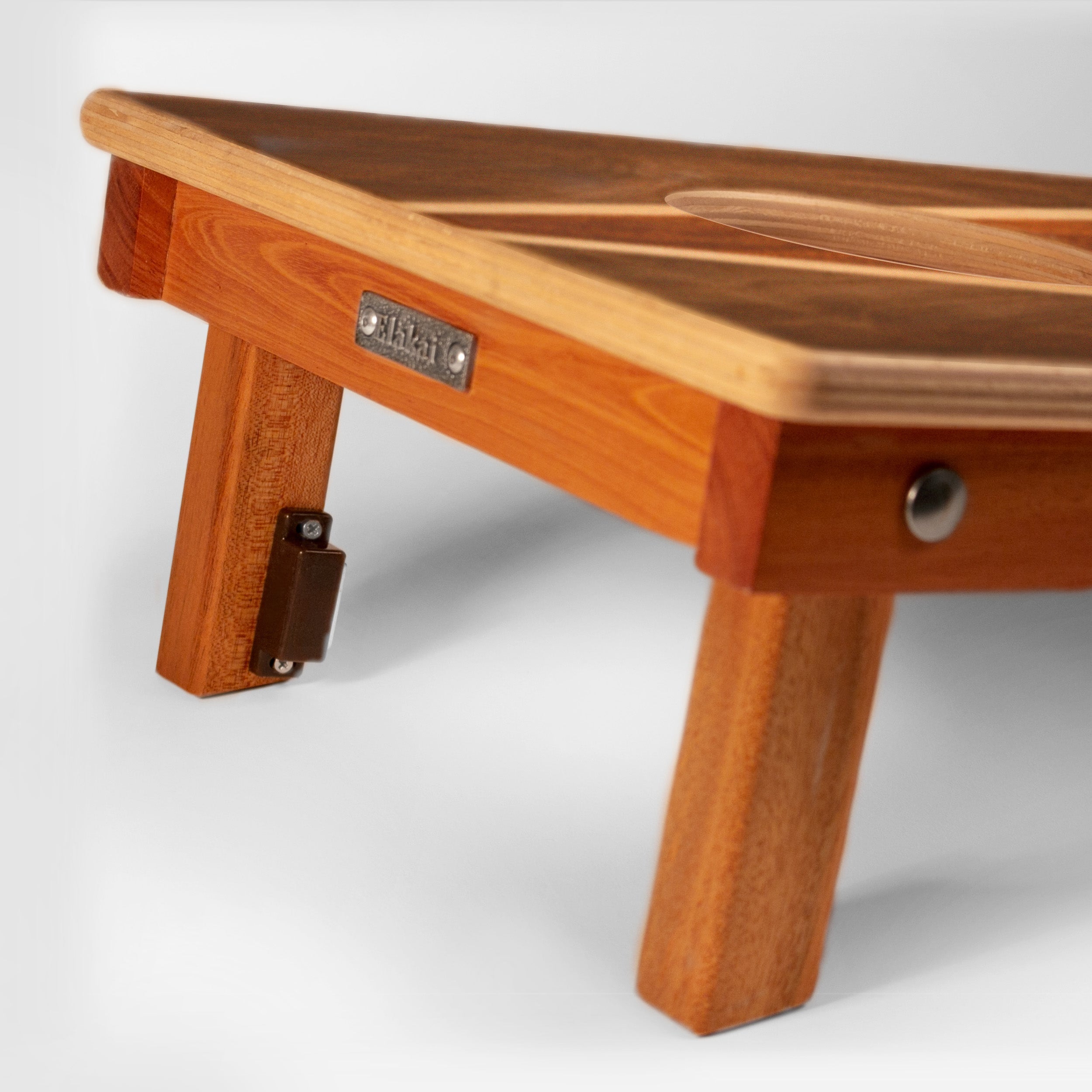 Close-up of a wooden folding table or bench with warm medium-brown wood grain, showing the tabletop, front frame, leg, and a small metal brand plate that reads KLAS.