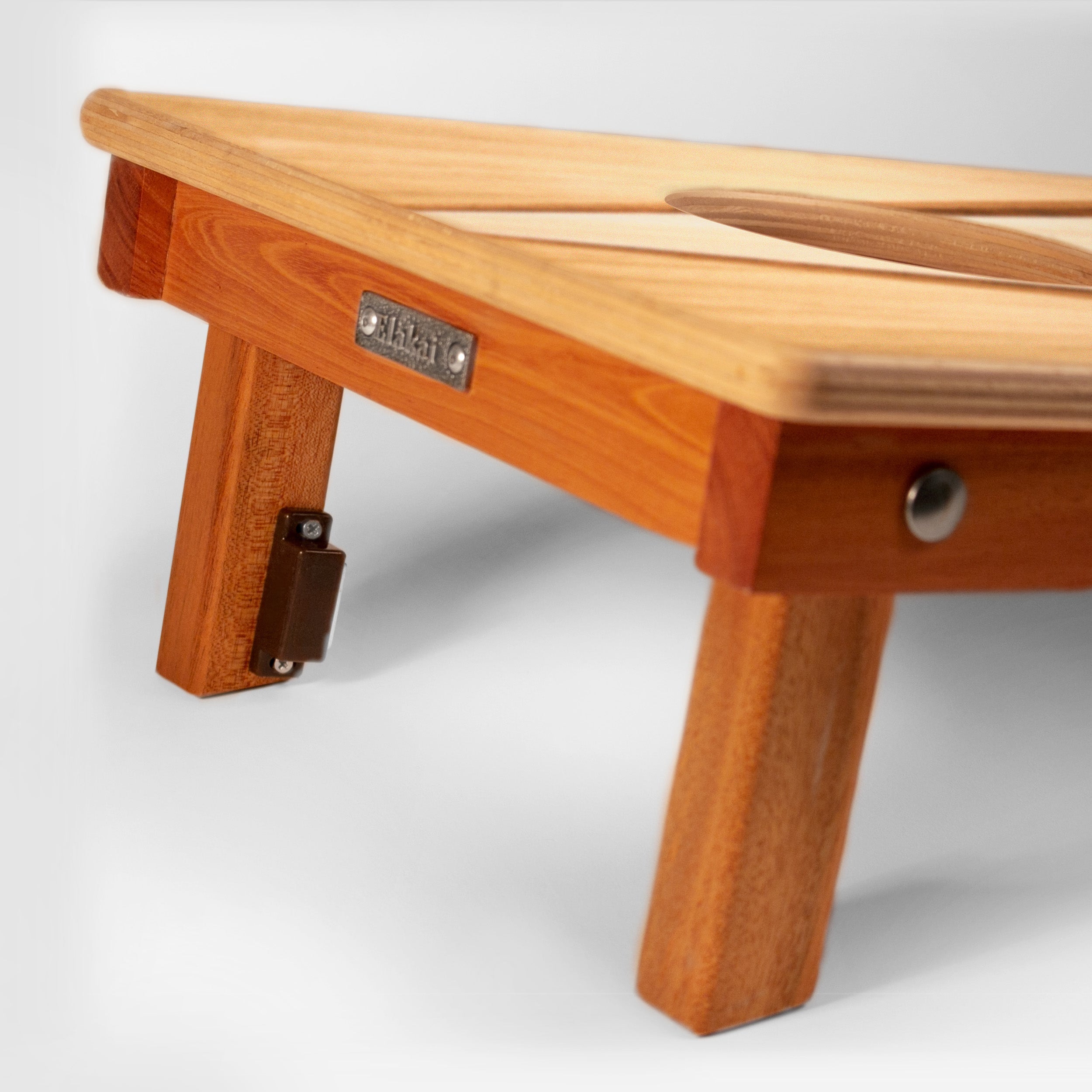 Close-up of a wooden folding table or bench with a warm honey-toned wood finish, showing the tabletop, leg, hinge, and a small metal brand plate that reads 'STUDY'.