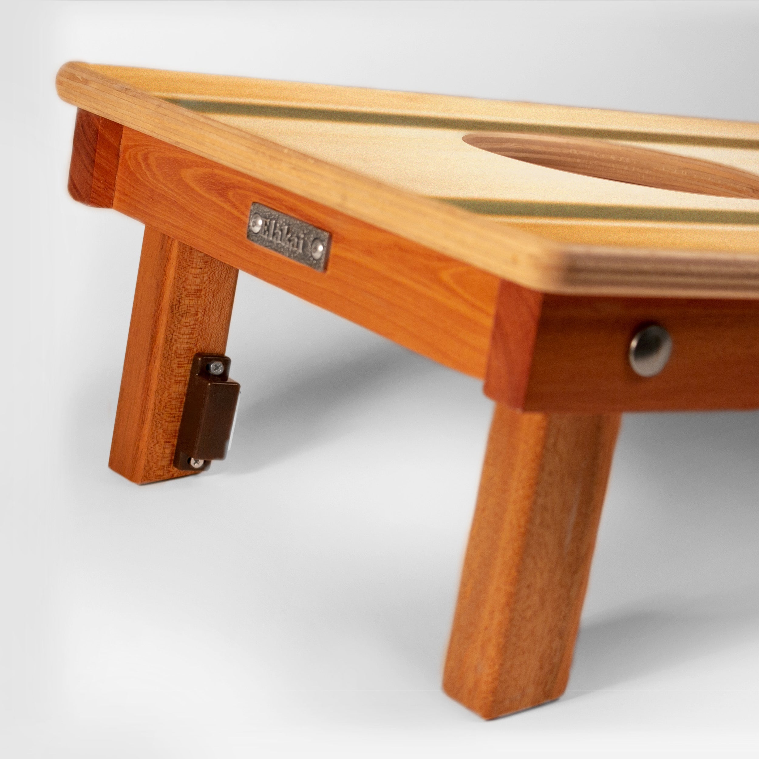Close-up of a wooden folding table corner with warm honey-toned wood legs, a light natural wood top, a metal brand plate reading 'STUDIES', and a hinge on the leg.