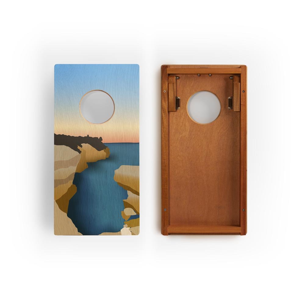 Two cornhole boards with a scenic design of a sunset over water and cliffs on one board, and a wooden texture on the other.