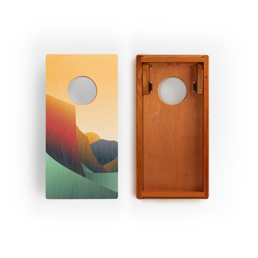 Two cornhole boards with one featuring a mountain landscape design on a white background