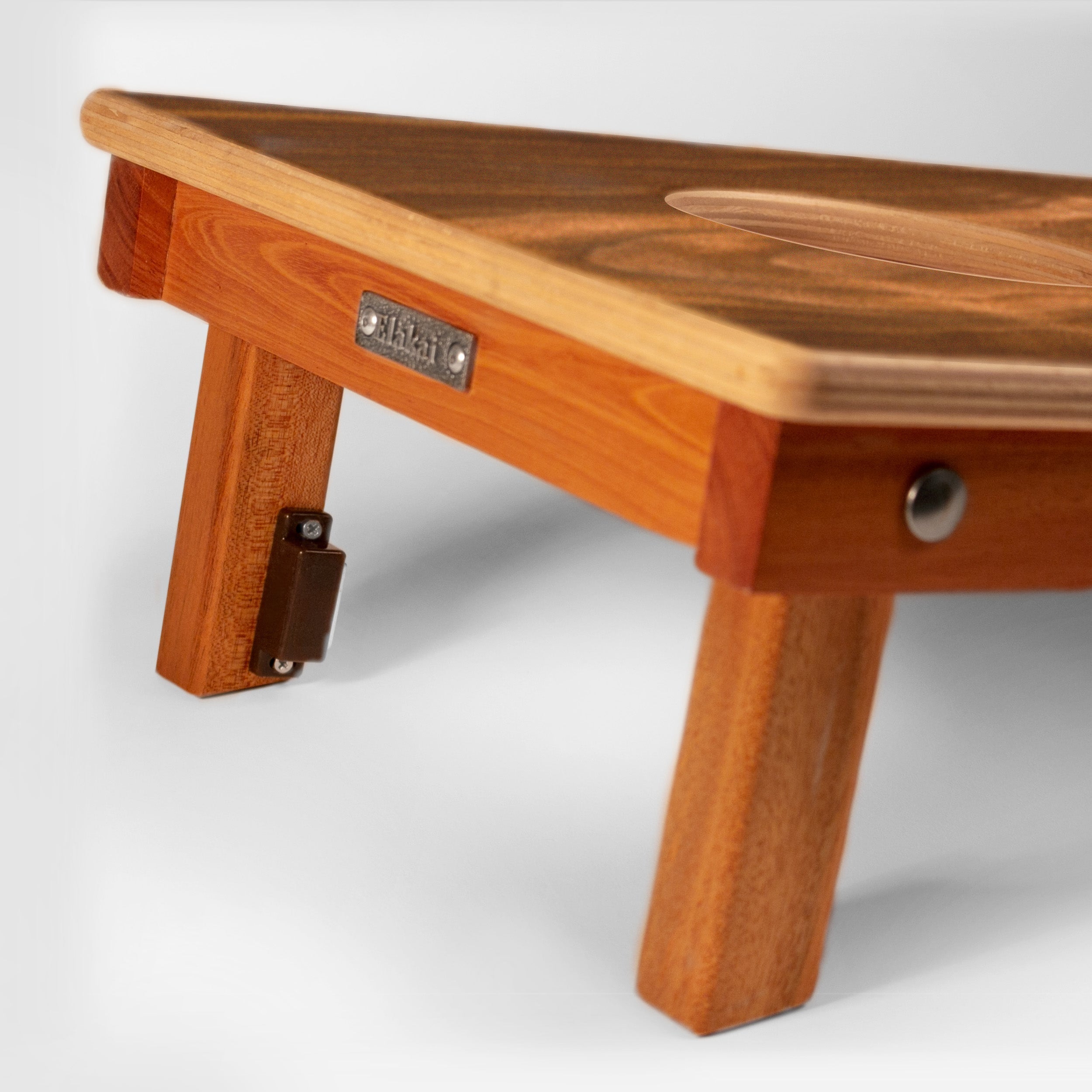 Close-up of a wooden folding table with warm medium-brown wood grain, showing the tabletop, leg, hinge, and a small metal brand plate that reads 'TIGER'.