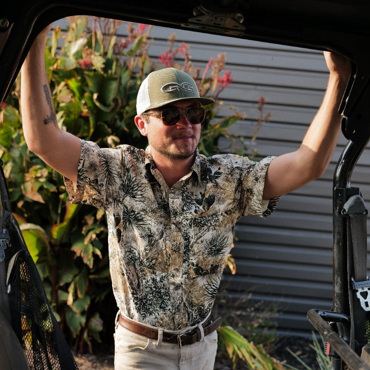 Man wearing a camouflage shirt and cap, standing next to an open vehicle with plants in the background.