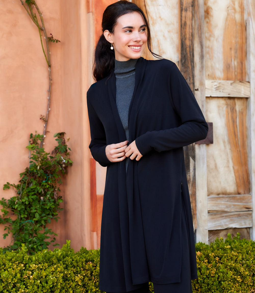 A woman wearing a long black open-front cardigan over a gray turtleneck sweater, standing outdoors in front of a rustic wooden door and terracotta wall.