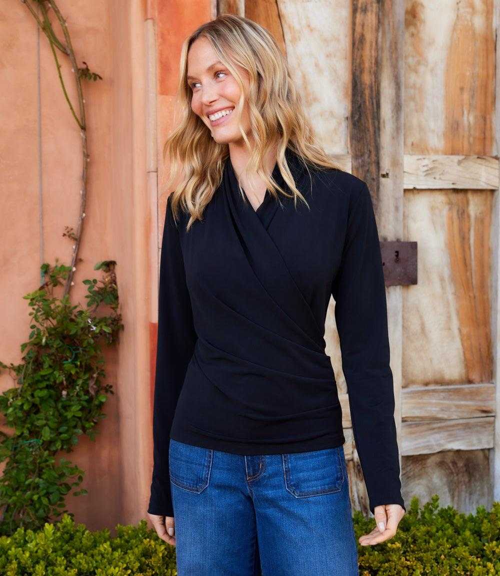 A woman wearing a black long-sleeve wrap-style top and blue jeans, standing outdoors in front of a rustic wooden door and terracotta wall.