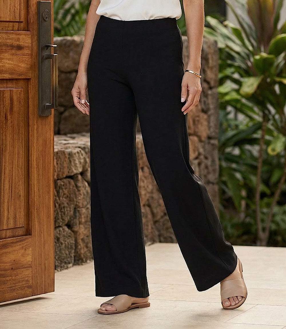 A person wearing black wide-leg pants with side cutouts, paired with a white top and beige slide sandals, standing in front of a wooden door and stone wall with green plants.