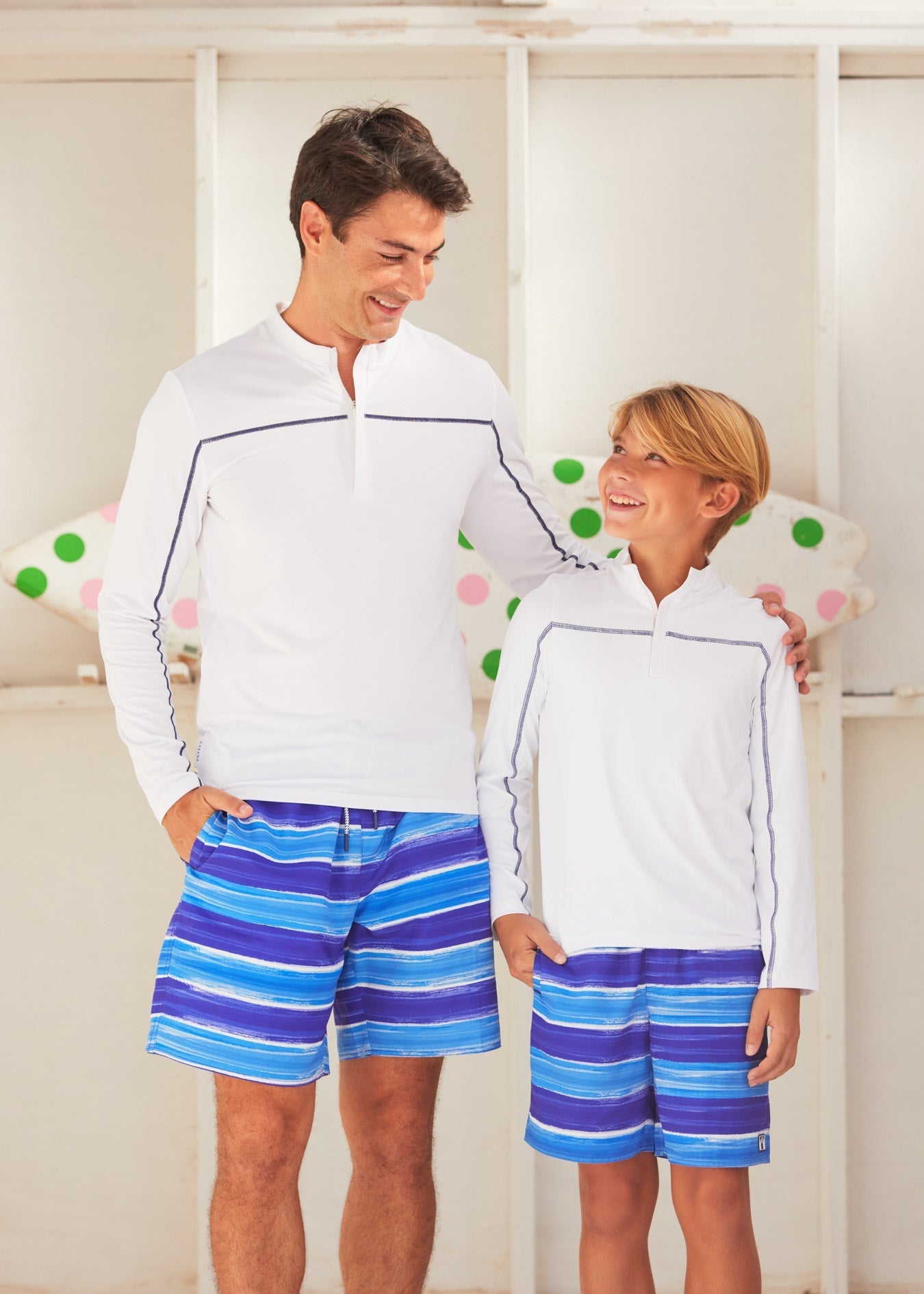 Man and woman wearing matching white hoodies and blue striped shorts standing in front of a polka dot curtain.