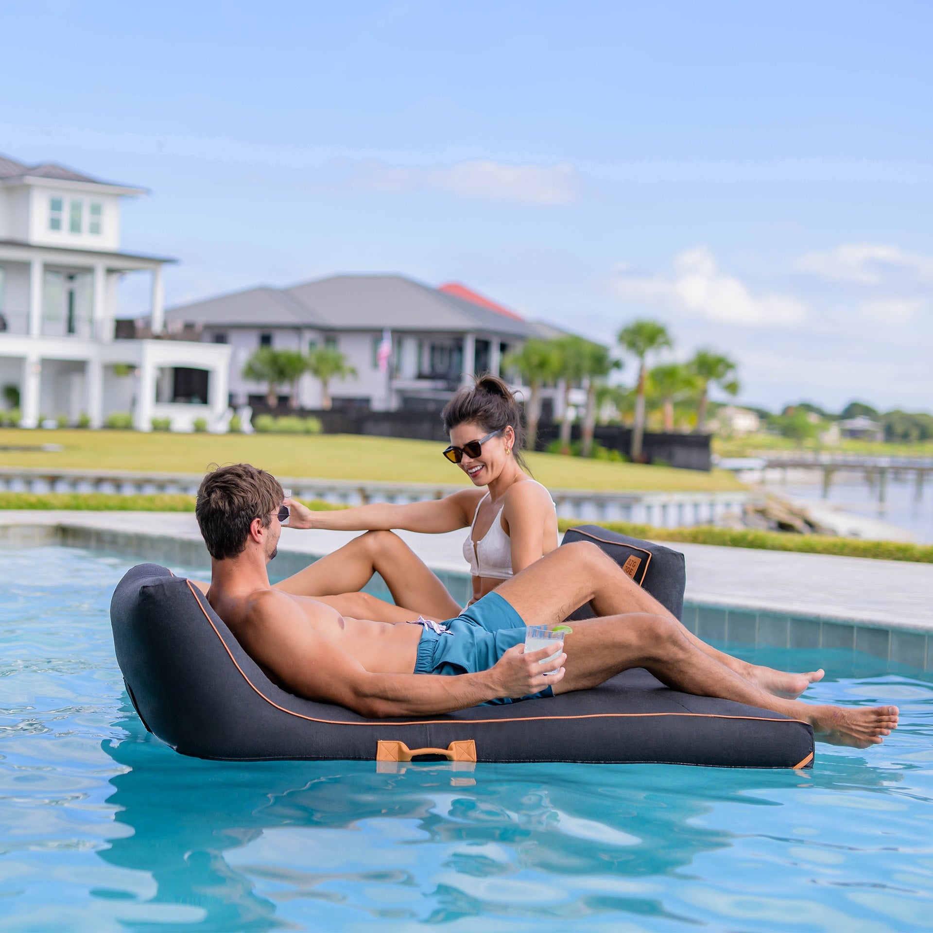 Two people relaxing on a floating inflatable lounger by a pool with a scenic background.