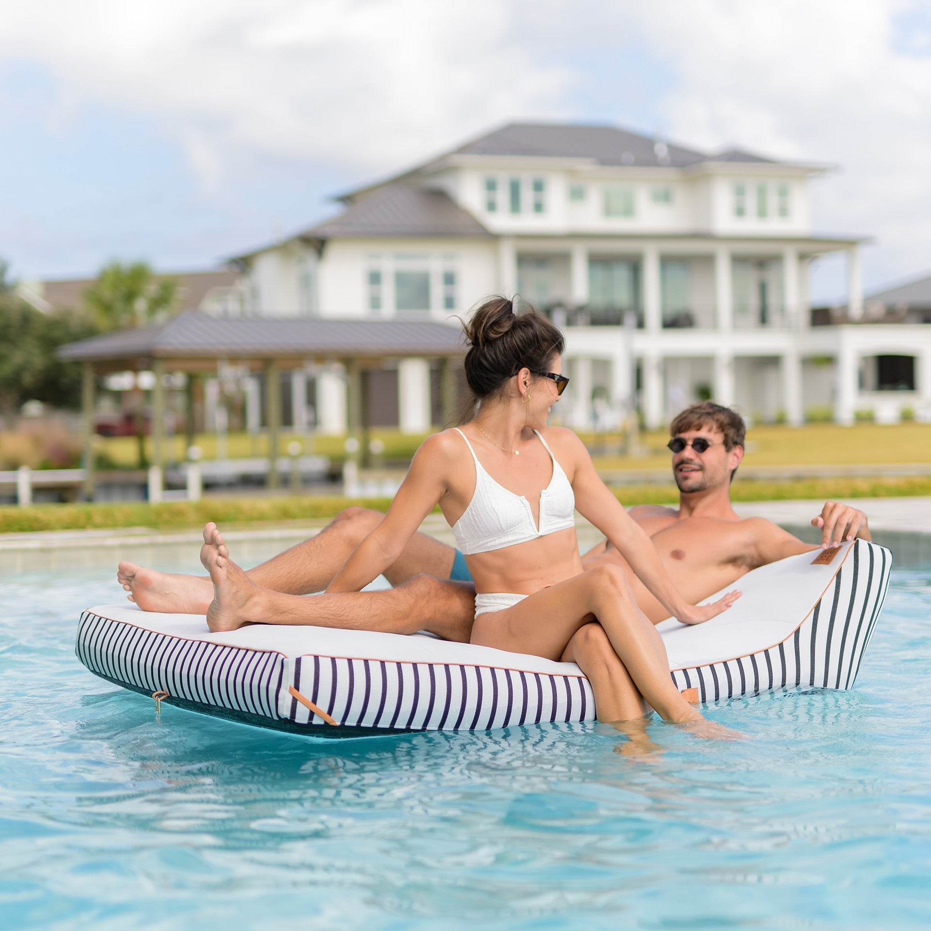 Two people relaxing on a striped inflatable pool float in a pool with a house in the background.