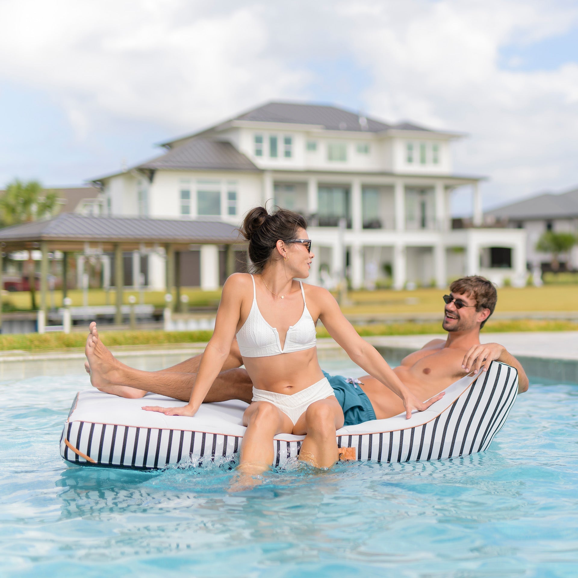 Two people sitting on a striped pool float in a swimming pool with a house in the background.
