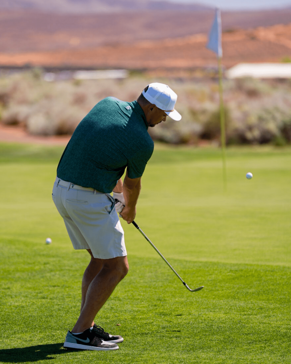 A male golfer in a dark green polo shirt, white shorts, a white cap, and black Nike golf shoes swings an iron on a golf course green, with a flag and desert hills in the background.