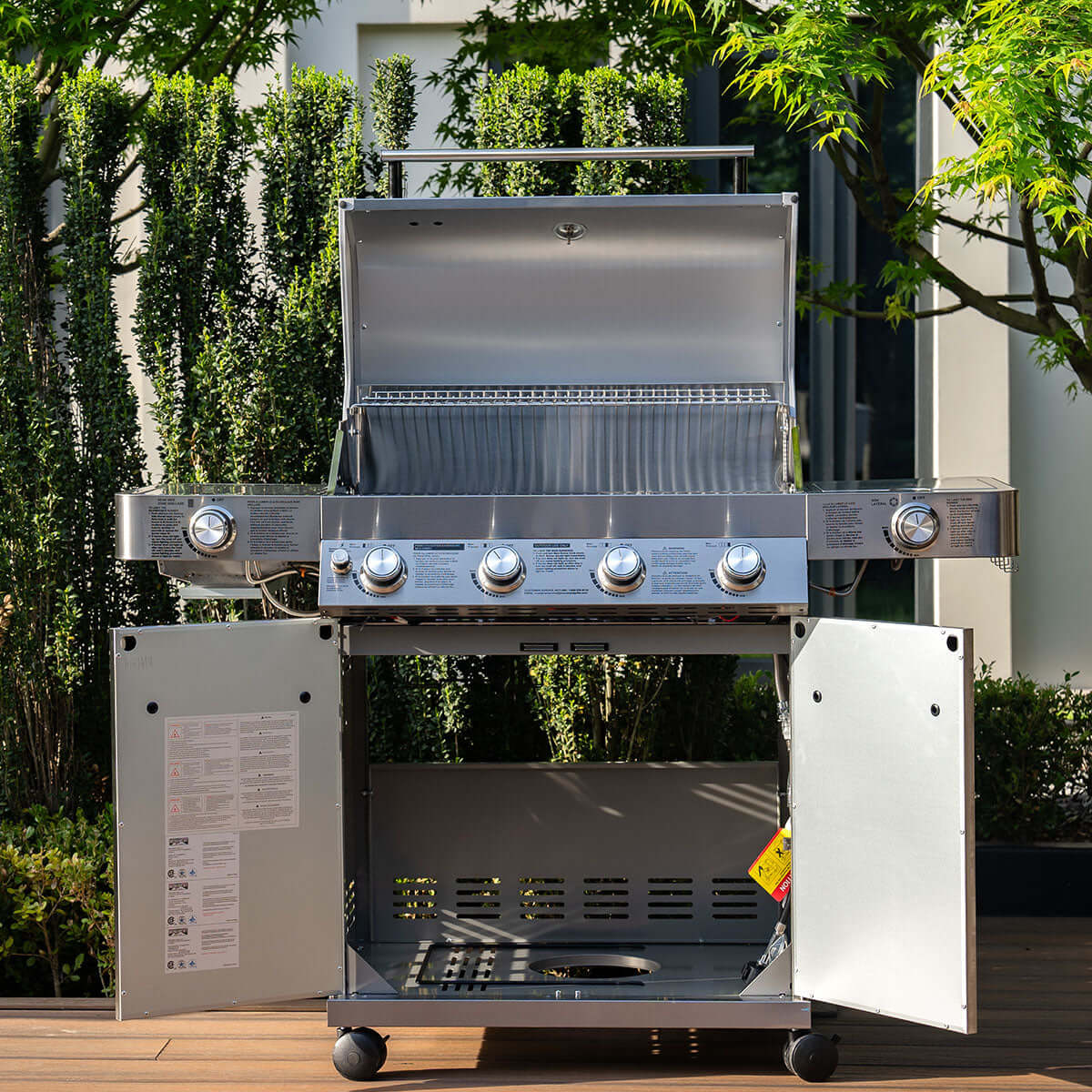 Outdoor grill with open side panels on a wooden deck with greenery in the background