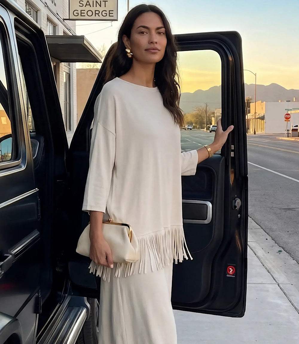 A woman stands by an open black vehicle door on a city street at sunset, wearing a cream-colored three-quarter sleeve top and matching pants with fringe trim, holding a small cream clutch.