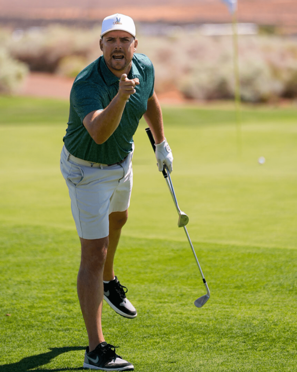 A man on a golf course wearing a dark green polo shirt, white shorts, a white cap, and black-and-white golf shoes, holding a golf club and pointing toward the camera.
