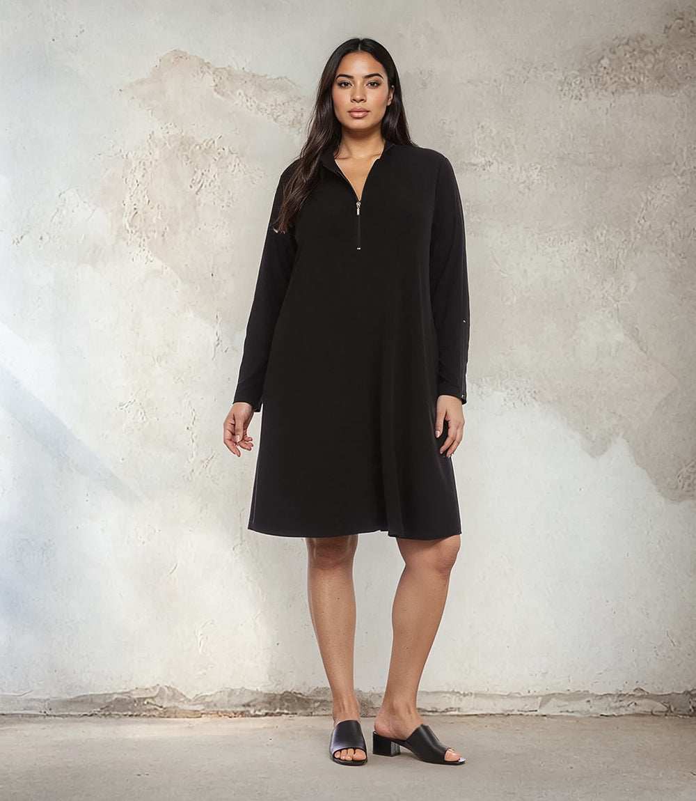 A woman modeling a black long-sleeve shift dress with a half-zip neckline, standing against a textured concrete wall.