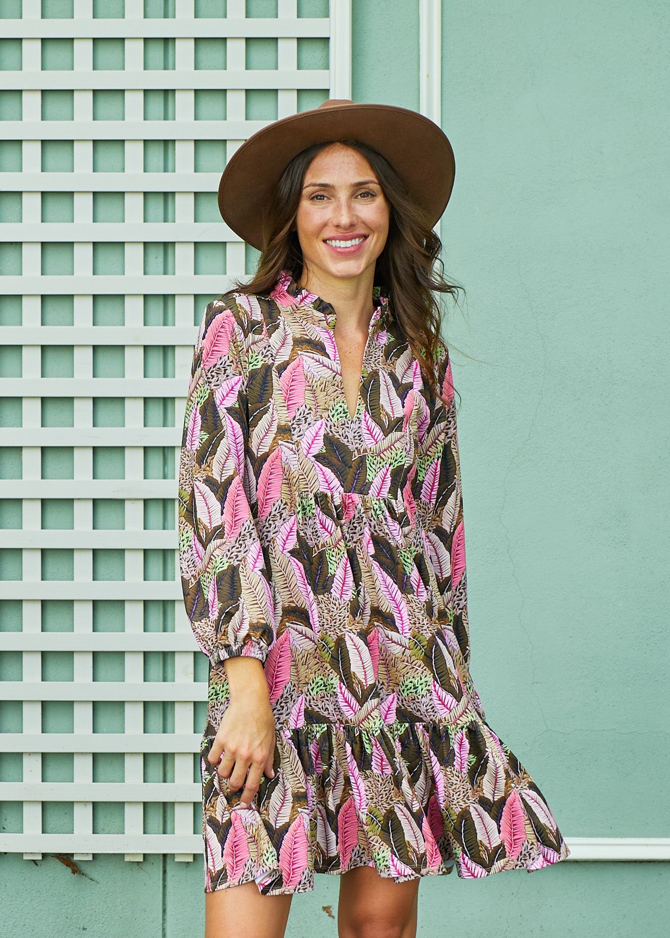 Woman wearing a patterned dress and hat against a light green wall.