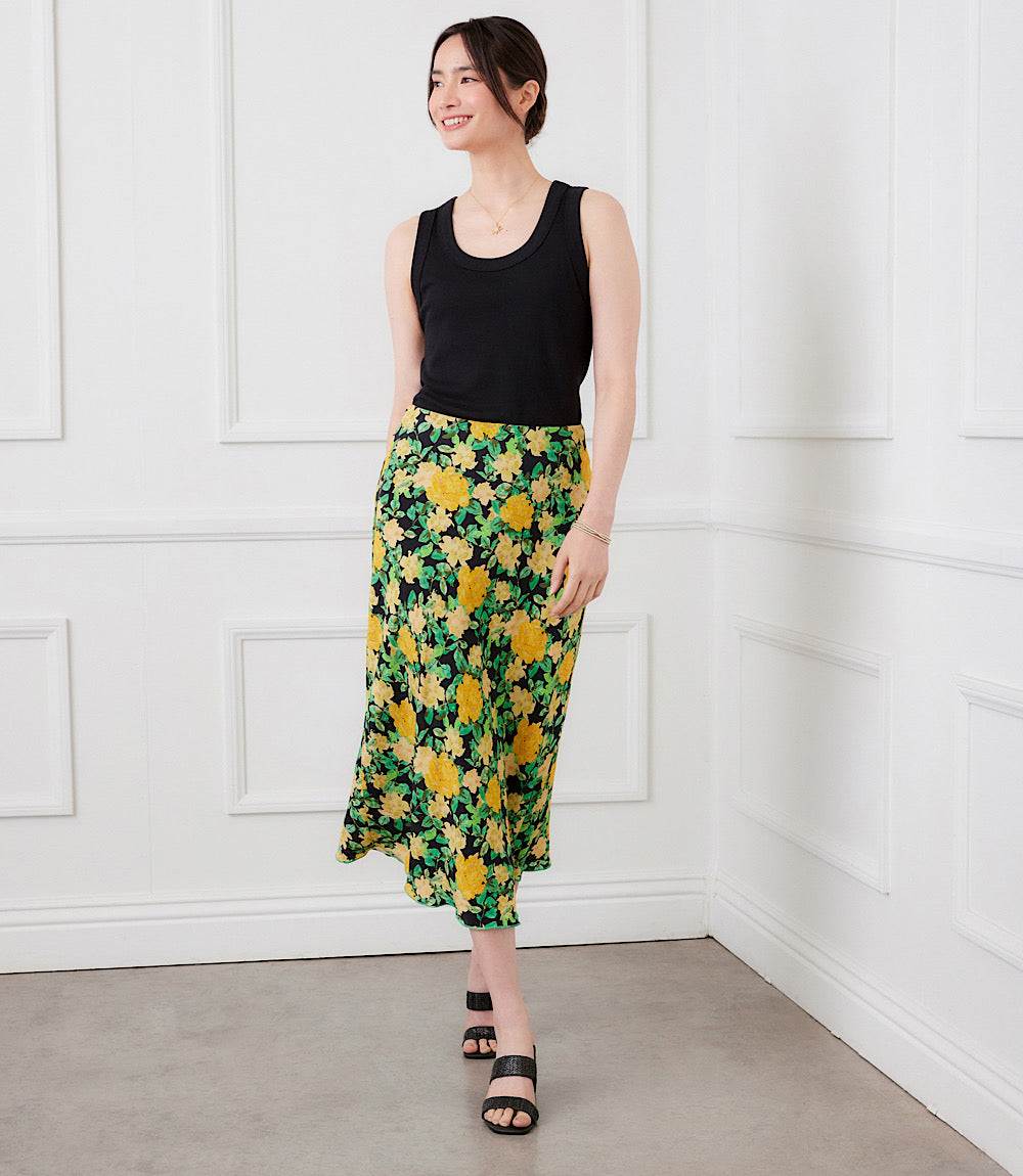 A woman stands in a white paneled room wearing a sleeveless black top and a green midi skirt with a yellow and white floral print. She is wearing black slide sandals.