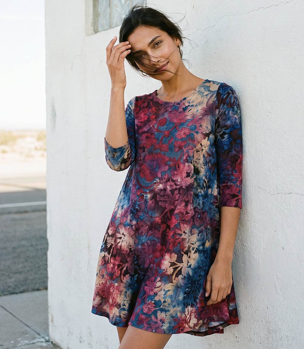 A woman wearing a short-sleeve, knee-length A-line dress with a vibrant floral print in shades of blue, magenta, and purple, leaning against a white stucco wall.