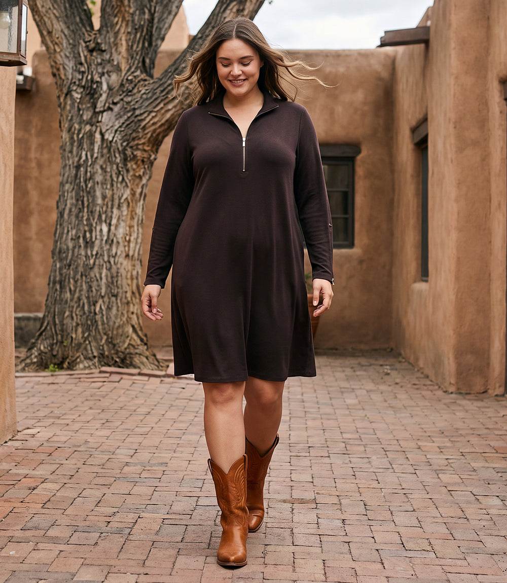 A woman wearing a dark brown long-sleeve half-zip dress walks outdoors on a brick path between adobe-style buildings and a large tree.