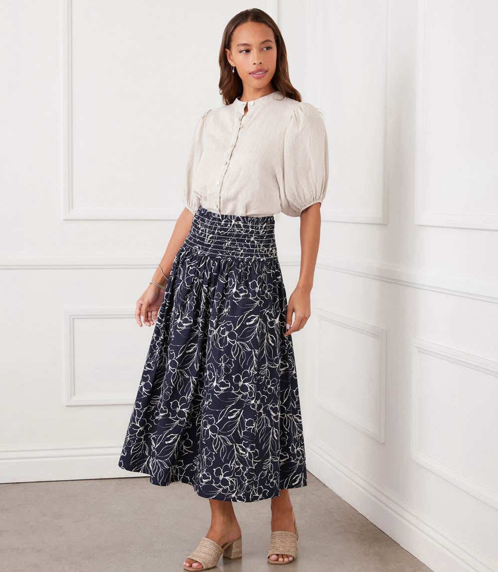 A woman modeling a white short-sleeve blouse and a navy midi skirt with a white floral print, standing in a white paneled room.
