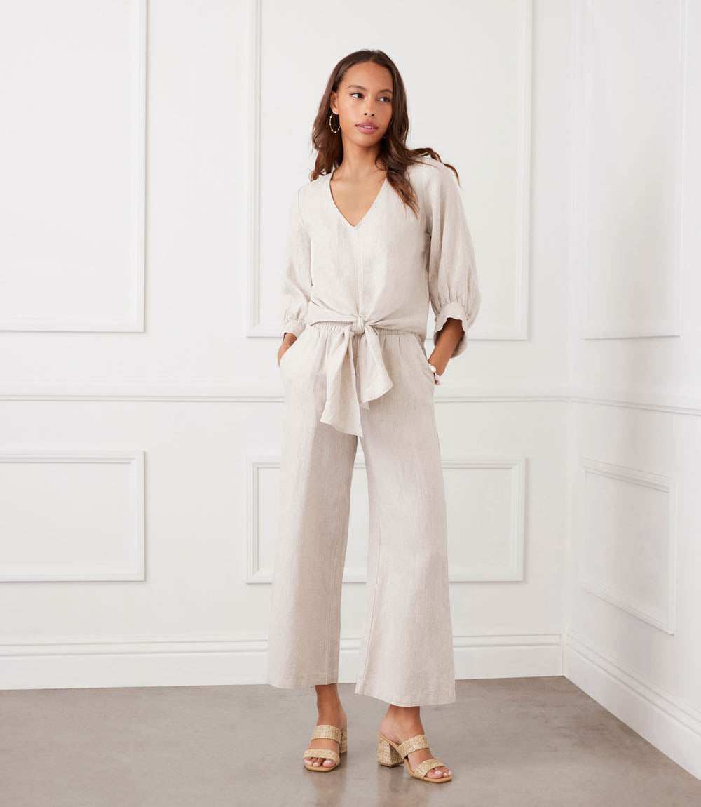 Woman wearing a beige jumpsuit standing against a white paneled wall.