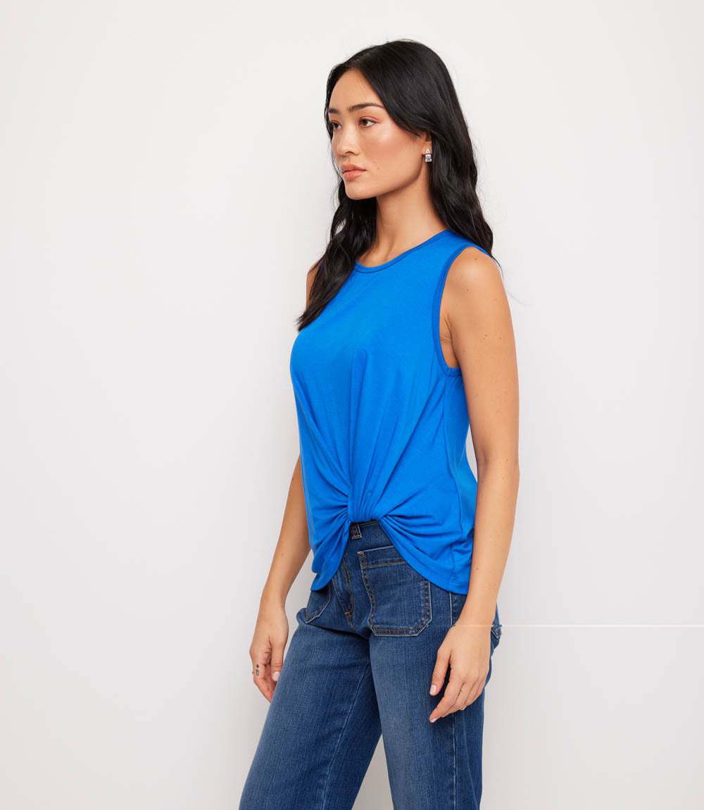 A woman modeling a bright blue sleeveless top with a twisted knot detail at the hem, paired with blue jeans, against a plain white studio background.