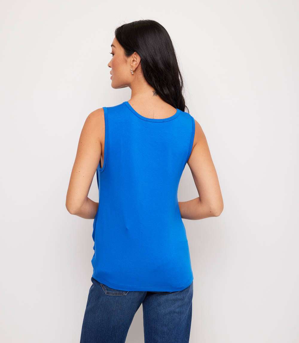 A woman shown from the back wearing a bright blue sleeveless tank top and dark blue jeans, standing against a plain light gray studio background.
