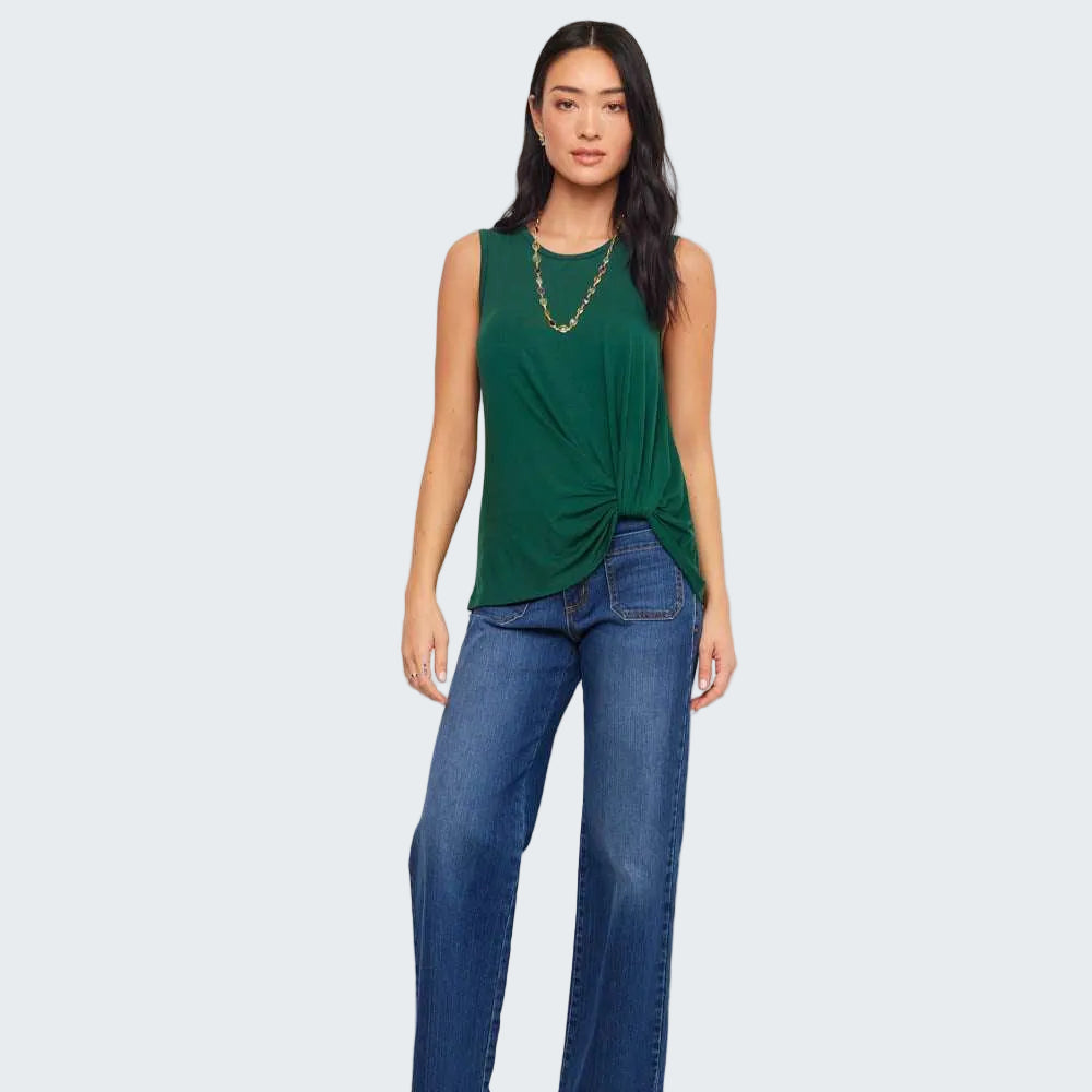 A woman modeling a sleeveless dark green top with a twisted front detail, paired with blue jeans and black sandals, standing against a plain white studio background.