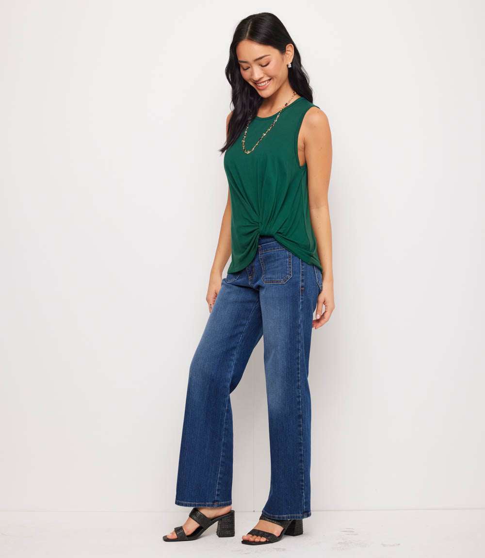 A woman modeling a sleeveless dark green top with a twisted knot detail at the front, paired with wide-leg blue jeans and black sandals, standing against a plain white studio background.