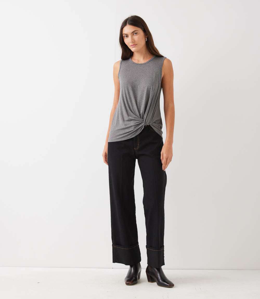 A woman modeling a gray sleeveless top with a twisted knot detail at the hem, paired with dark wide-leg pants and black ankle boots, against a plain white studio background.