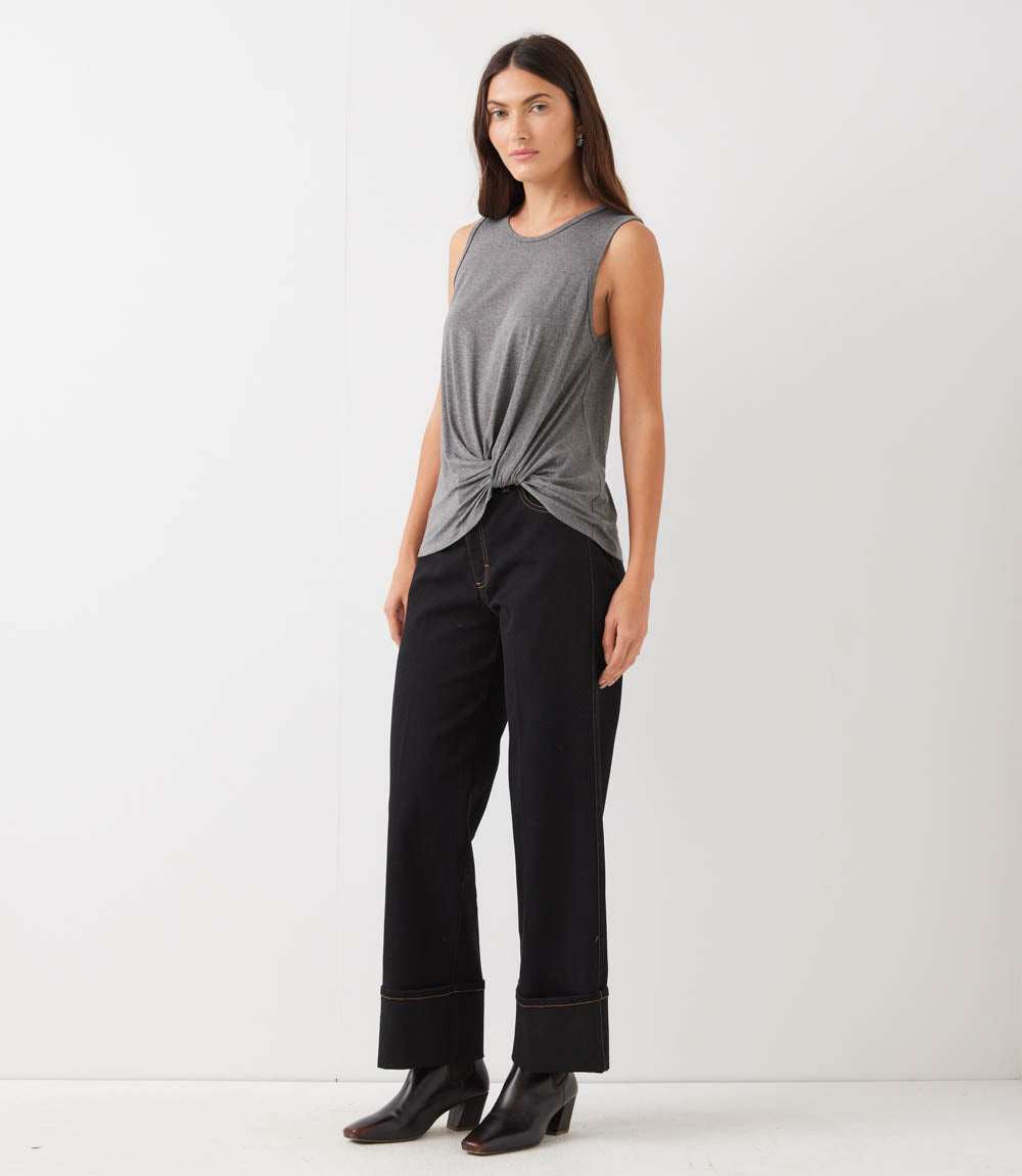 A woman modeling a sleeveless gray knit top with a twisted knot detail at the front, paired with black wide-leg jeans and black heeled boots, against a plain white studio background.