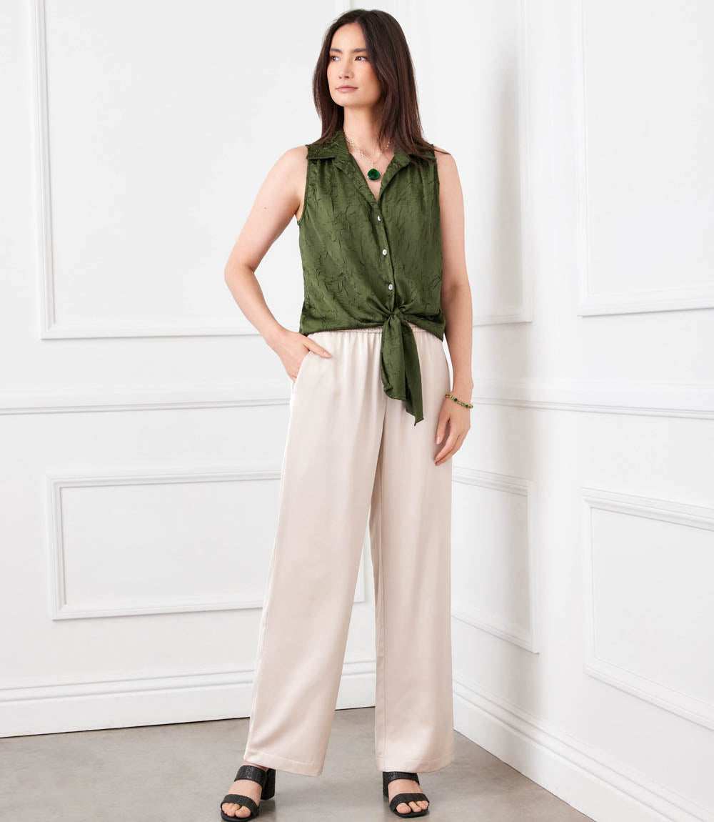 Woman wearing a green sleeveless shirt and beige pants against a white wall.