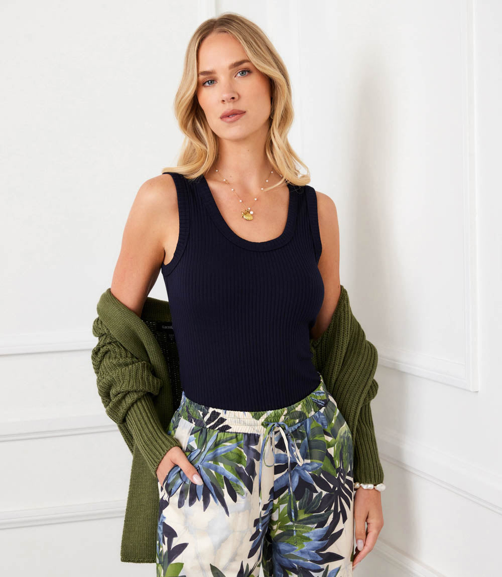 A woman stands against a white paneled wall wearing a navy ribbed tank top, an olive green cardigan draped off her shoulders, and cream drawstring pants with a blue tropical leaf print. She is also wearing layered gold necklaces.
