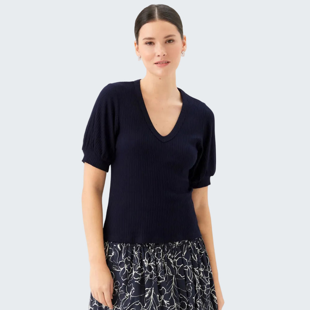 A woman modeling a navy blue ribbed short-sleeve V-neck knit top paired with a navy and white floral print skirt, set against a plain light gray studio background.