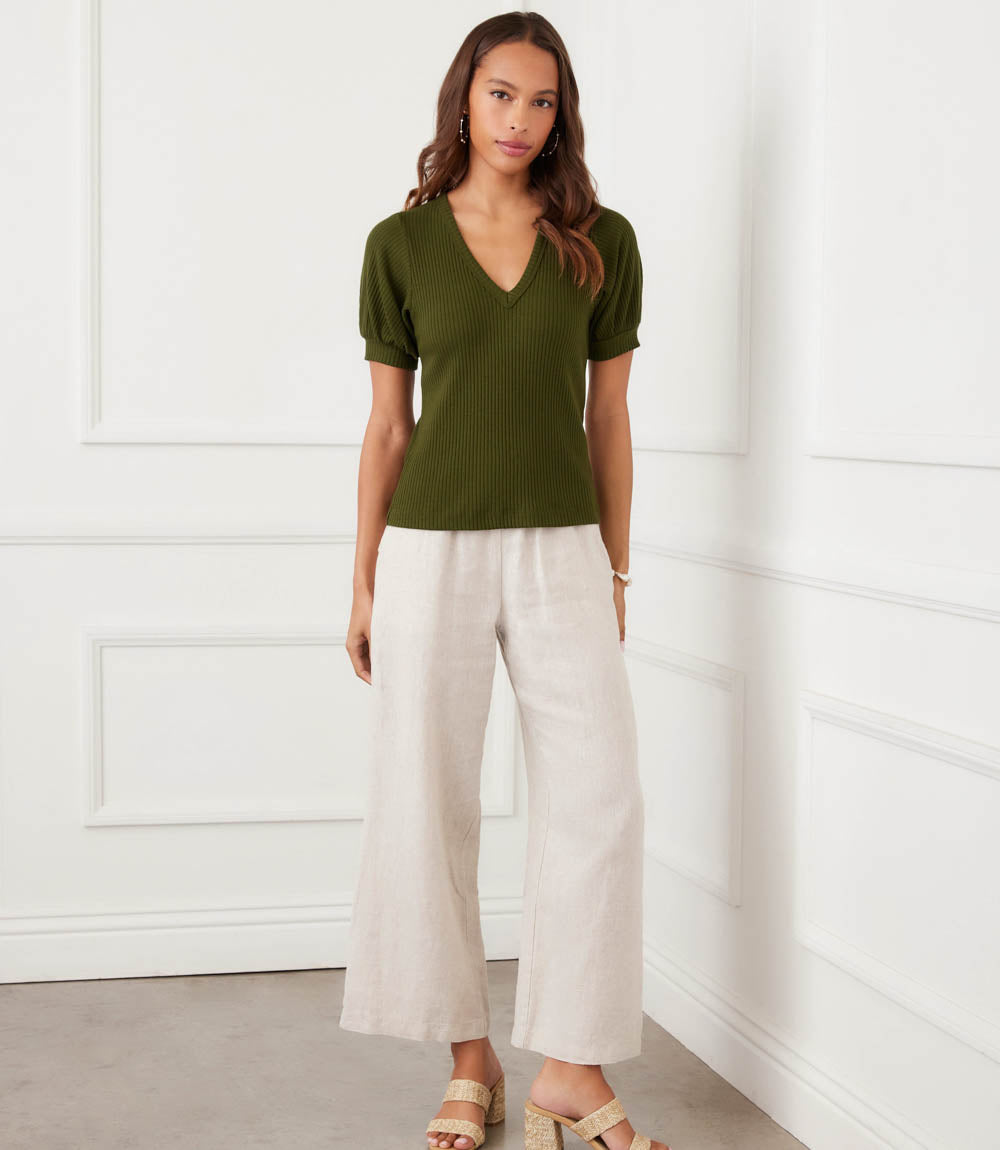 Woman wearing an olive green top and white pants against a white wall.