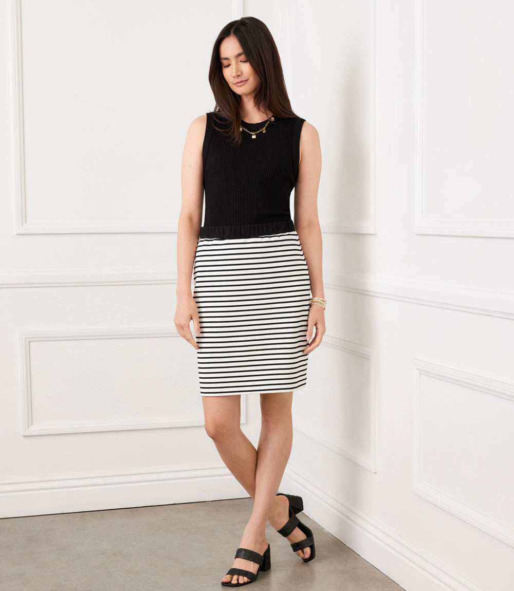 Woman wearing a black sleeveless top and black and white striped skirt against a white wall.