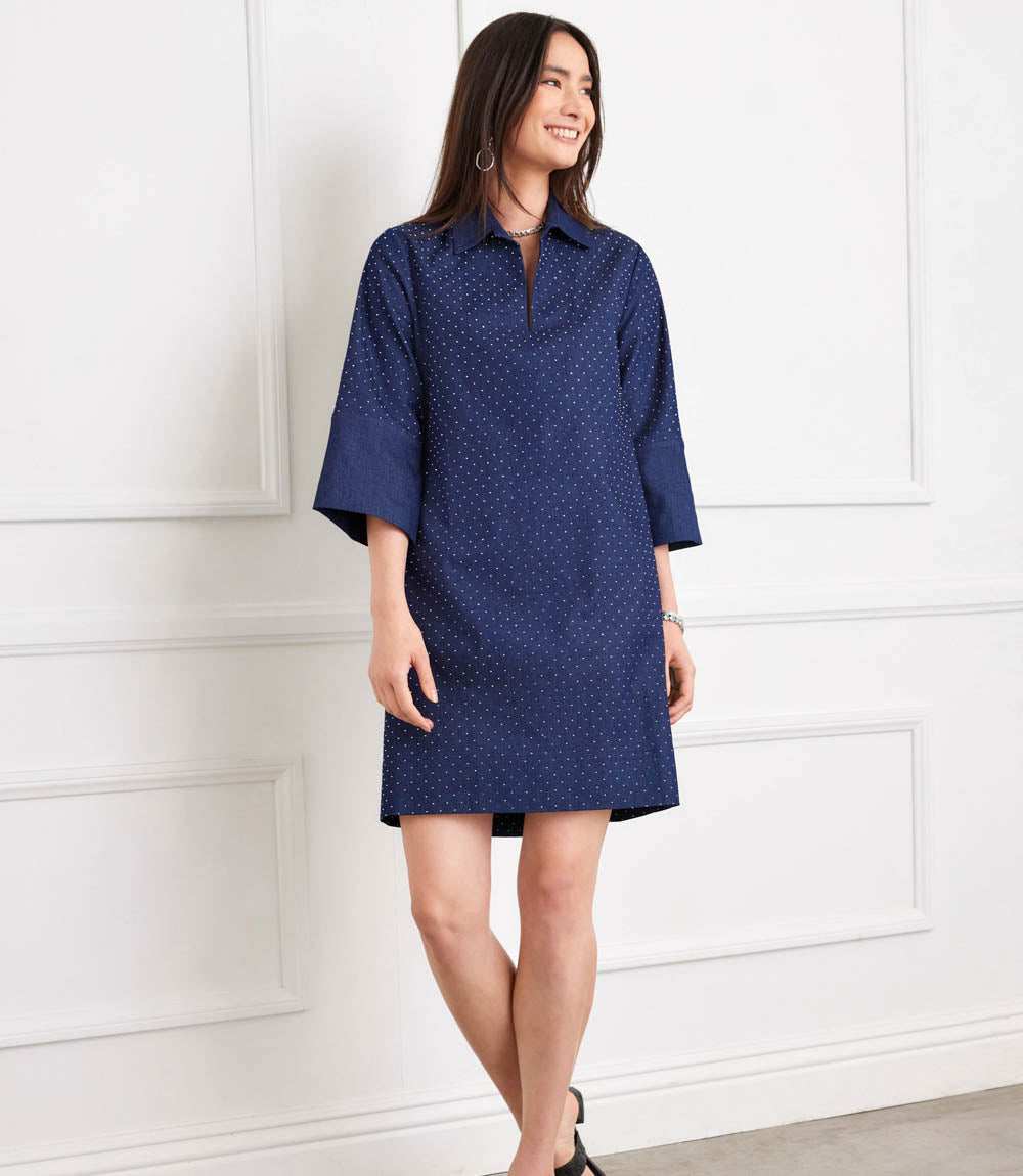 A woman wearing a navy blue short-sleeve collared dress with a small white polka dot pattern, standing against a white paneled wall.