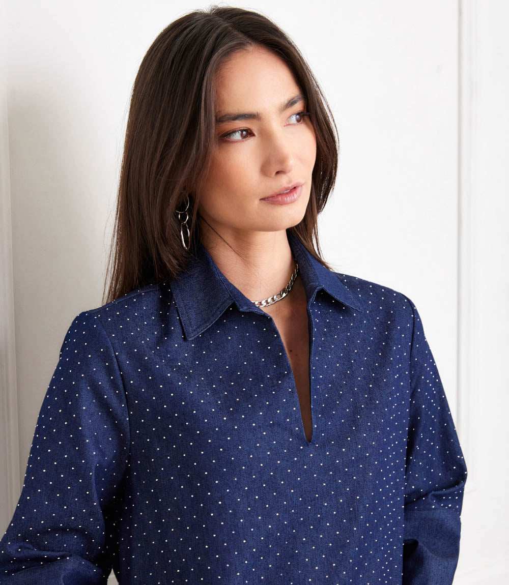 A woman wearing a navy blue long-sleeve button-down shirt with a small white polka dot pattern, styled with a silver chain necklace against a light off-white background.